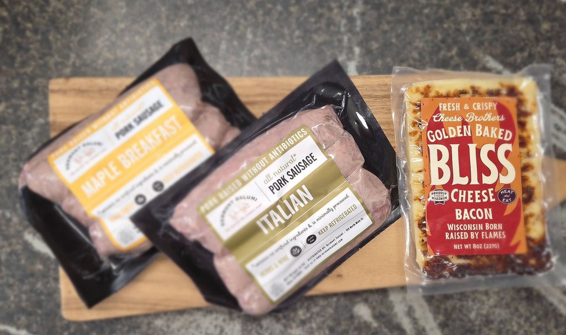 Three packaged food items—maple breakfast sausage, Italian pork sausage, and Golden Baked Bliss bacon cheese—on a board.