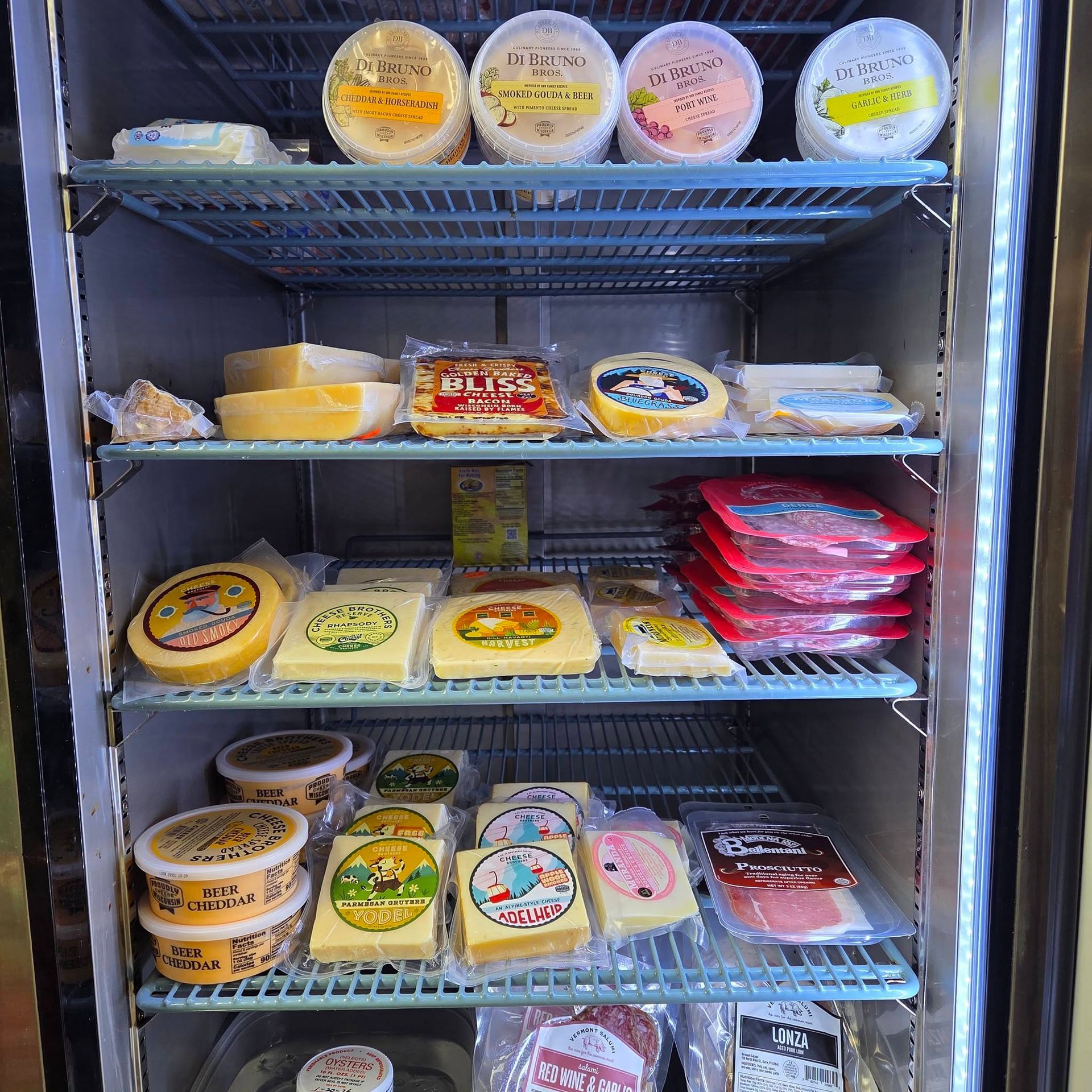 A refrigerated display case stocked with various types of cheese, meats, and deli items on metal shelves.