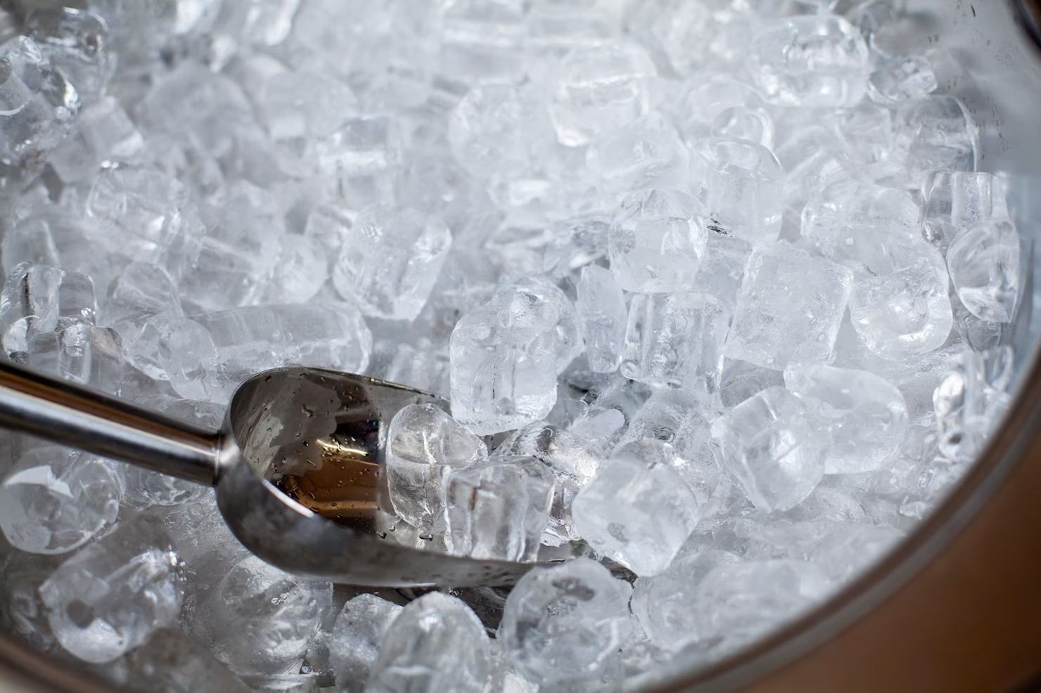 A metal scoop resting in a bucket filled with clear ice cubes.