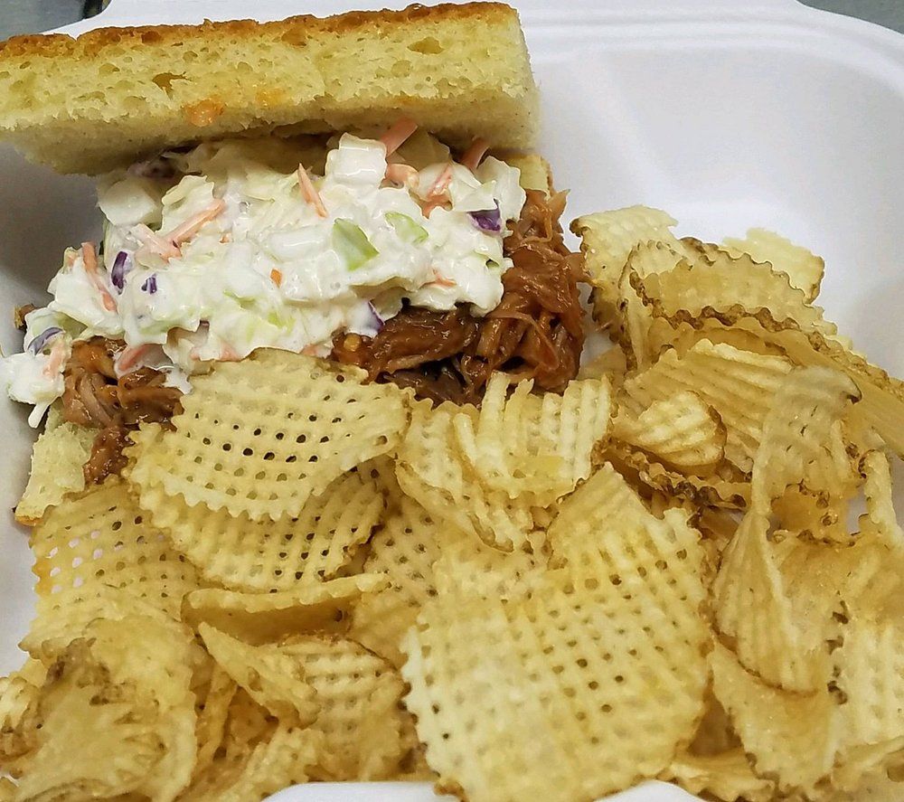 A pulled pork sandwich with coleslaw on cornbread, served with a side of waffle-cut potato chips in a white container.