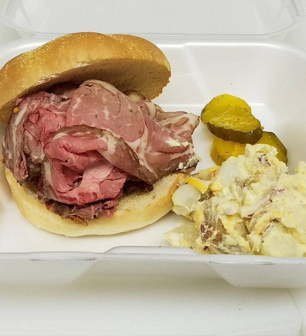 A roast beef sandwich on a bun served with a side of potato salad and pickle slices in a white styrofoam container.