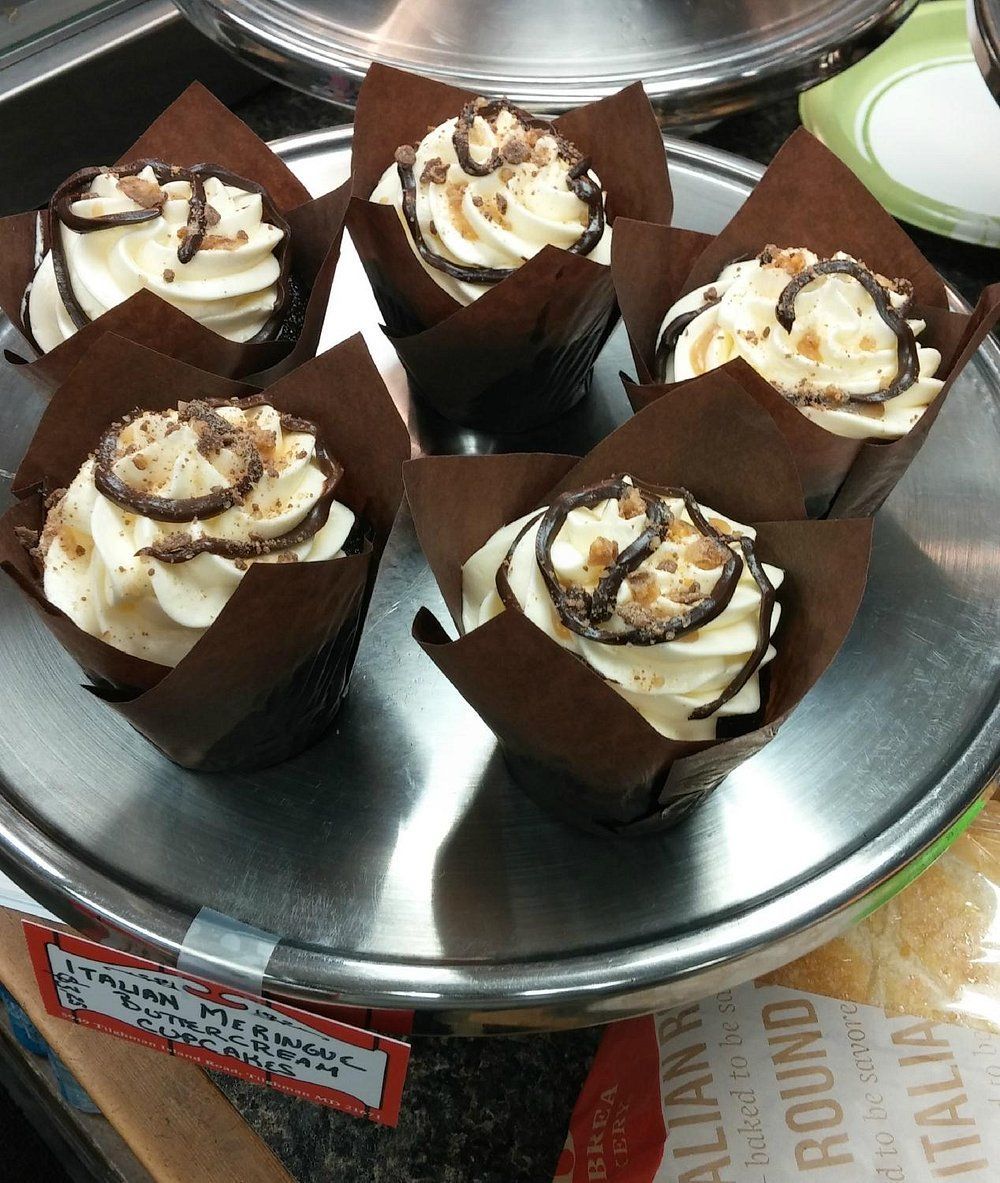 Five cupcakes with white frosting, chocolate drizzles, and sprinkles in brown paper liners, displayed on a metal stand.