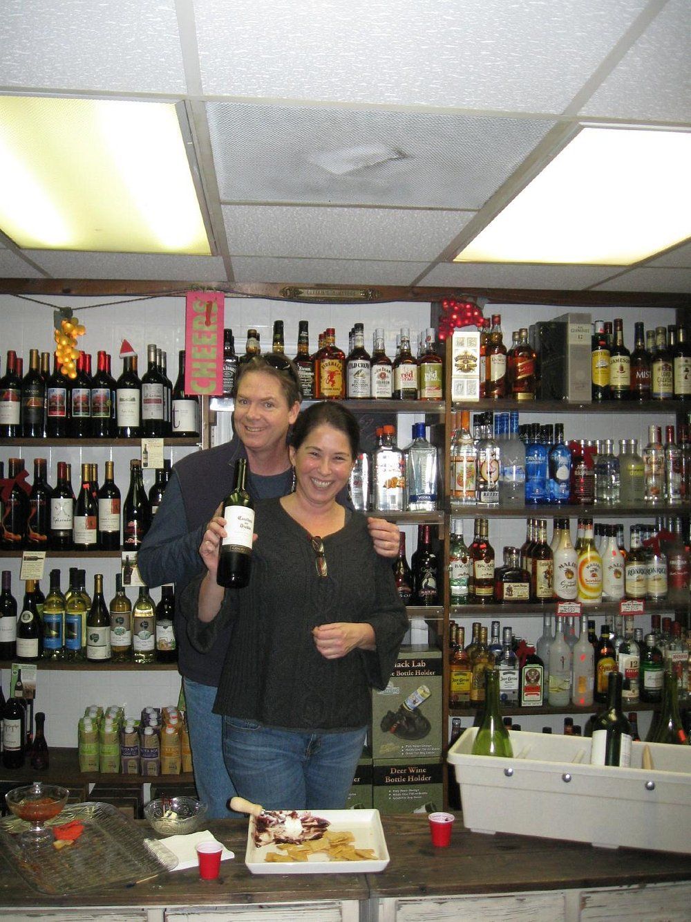 A smiling couple stands behind a bar filled with liquor bottles, one holding a wine bottle, with a plate of food in front.