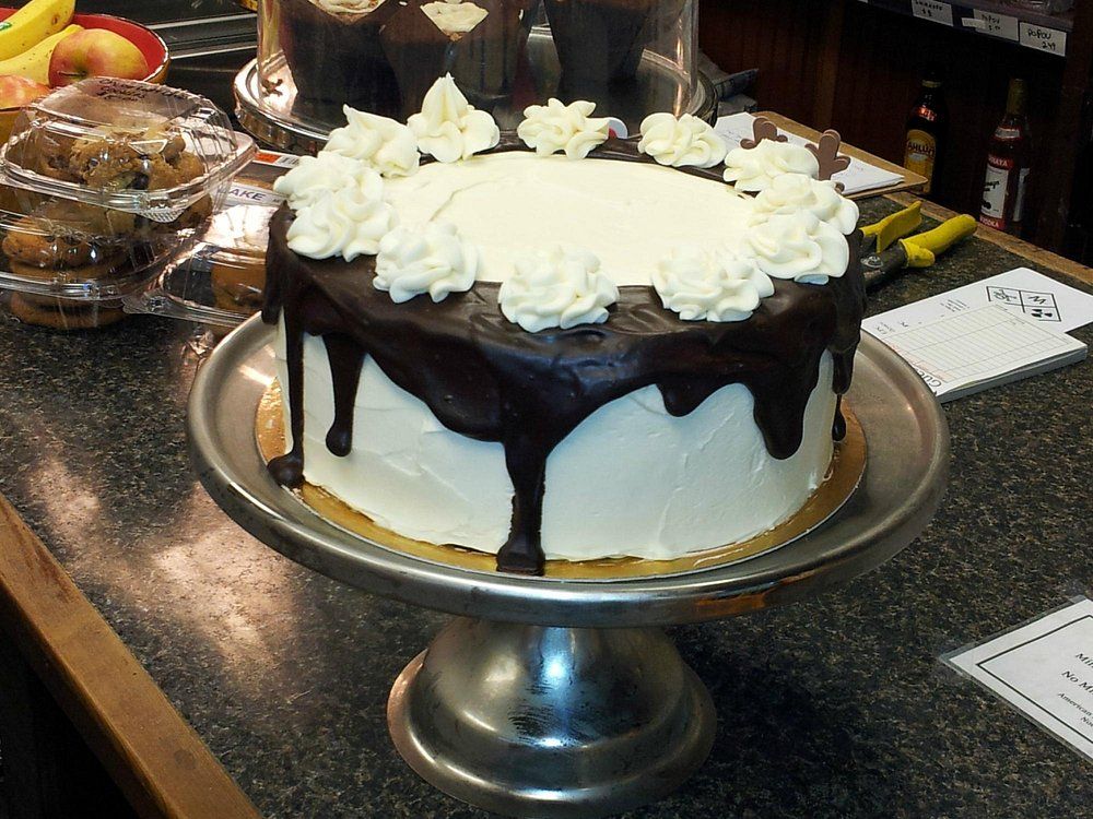 A white-frosted cake with a chocolate drip glaze and piped cream rosettes on top, displayed on a silver cake stand.