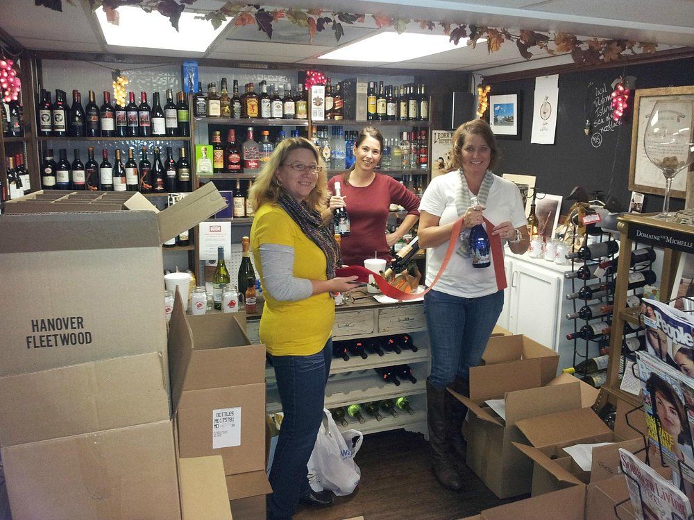 Three people stand behind a wine shop counter filled with bottles, some packing items into cardboard boxes.