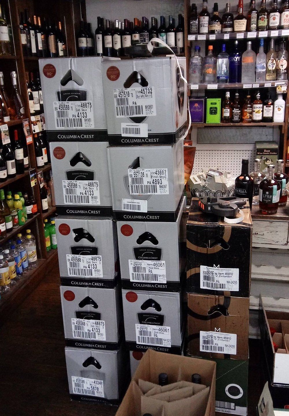 Stacks of cardboard liquor boxes with handwritten labels sit in an aisle of a liquor store, surrounded by full shelves.