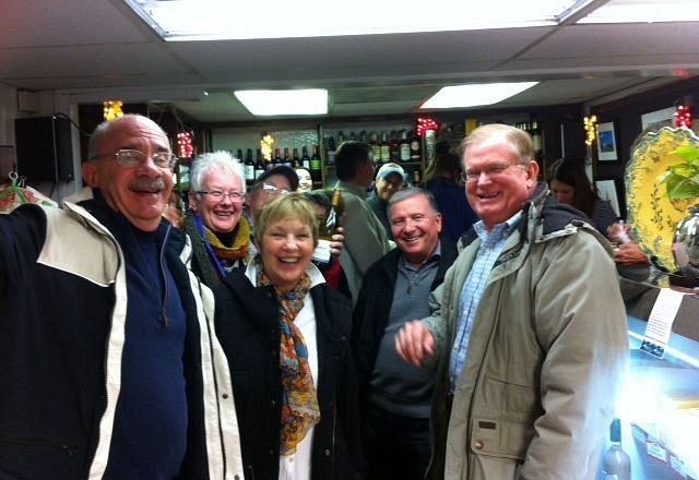 A group of five smiling individuals pose together in a warmly lit, casual indoor setting with shelves of bottles behind.