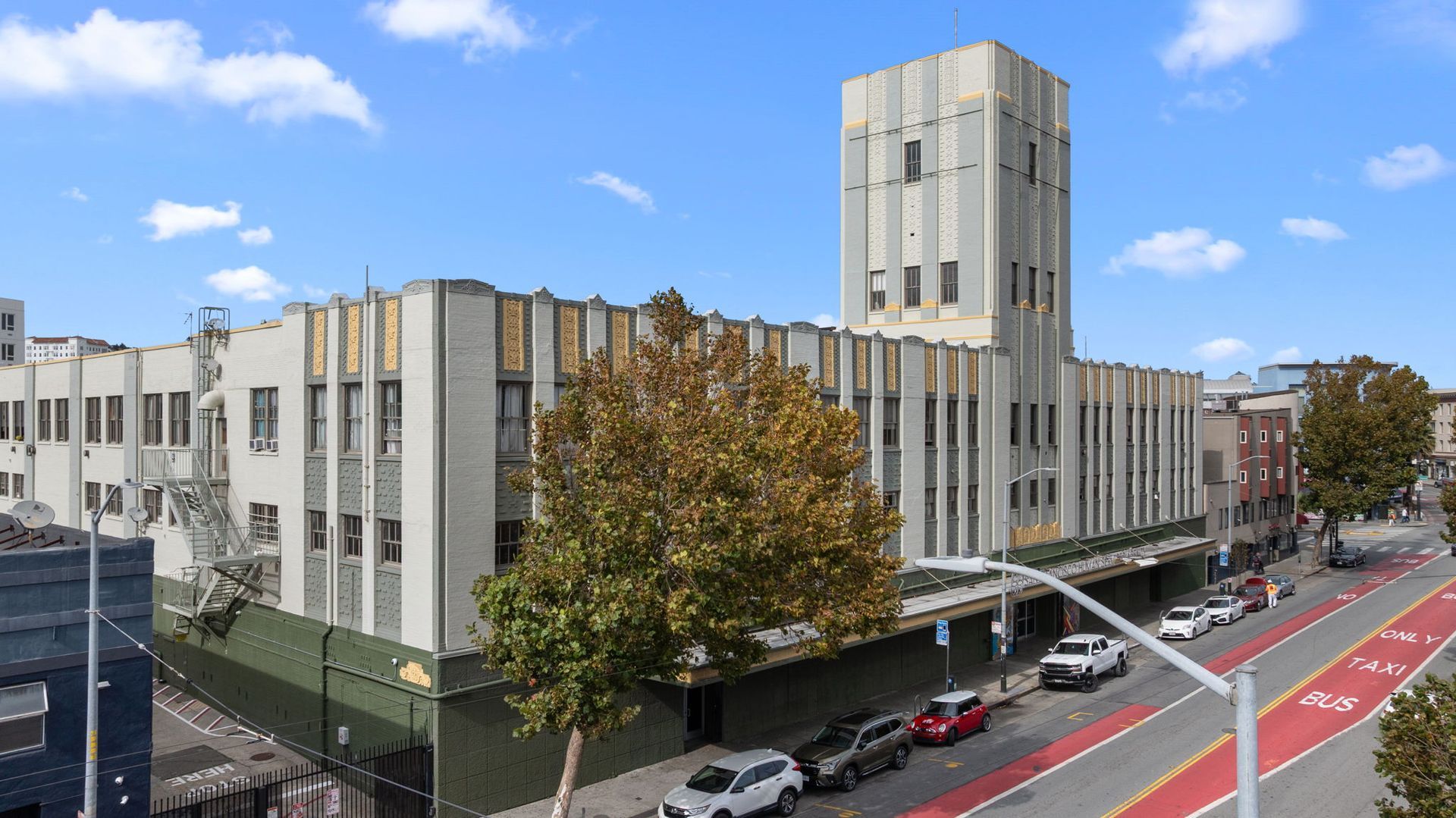Gray art deco building with a tower, street view. Yellowing tree in front, cars parked on the street.