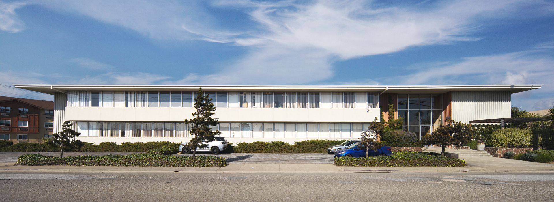 Building with a sign partially obscured by bushes, next to a street with cars and power lines.