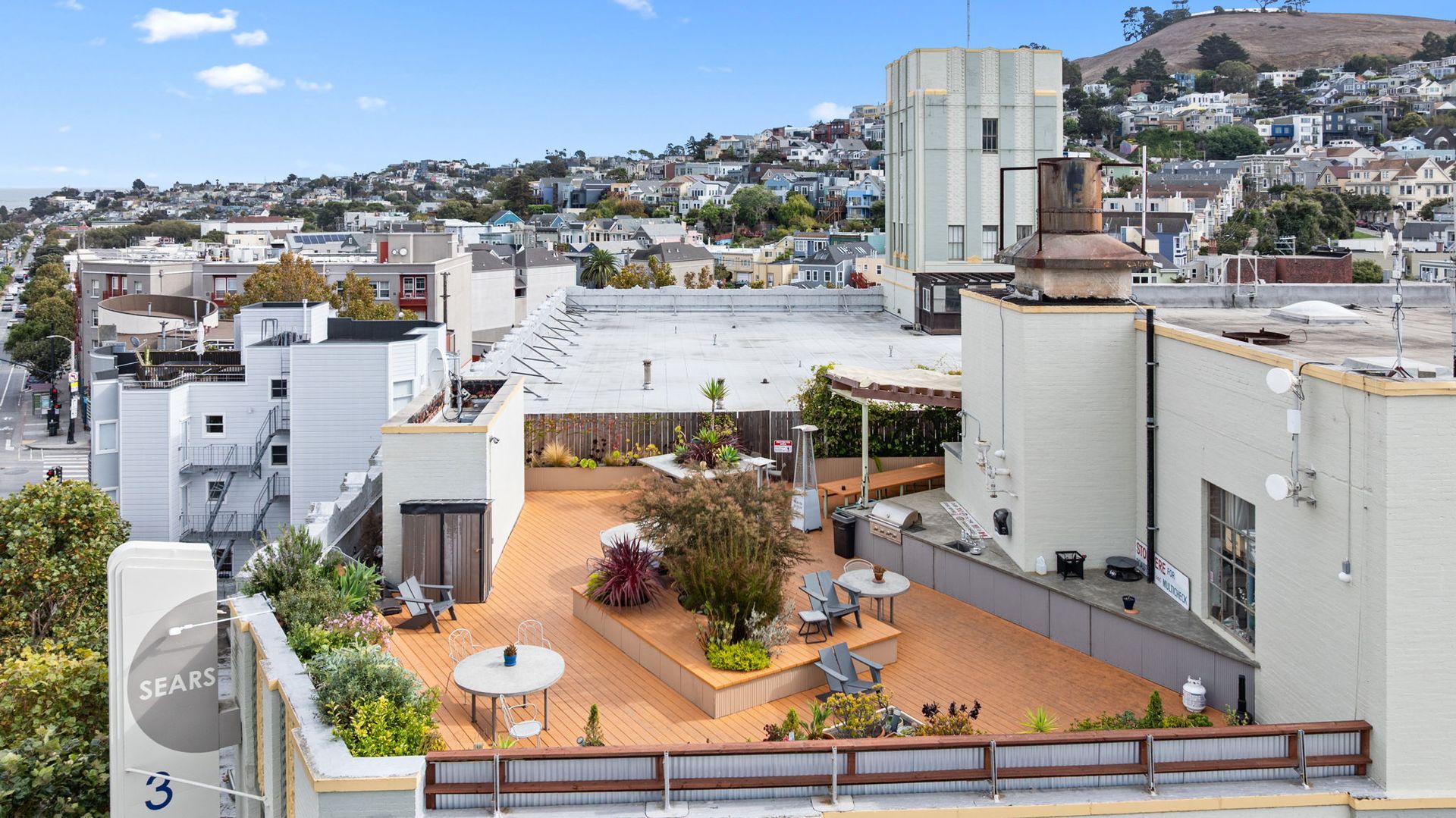 Rooftop patio with seating, plants, and city view.  Buildings, trees, and blue sky.