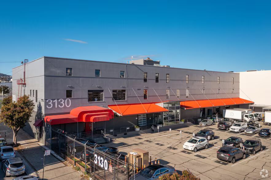 Gray industrial building with red awnings, a parking lot, and parked cars. Sign reads 