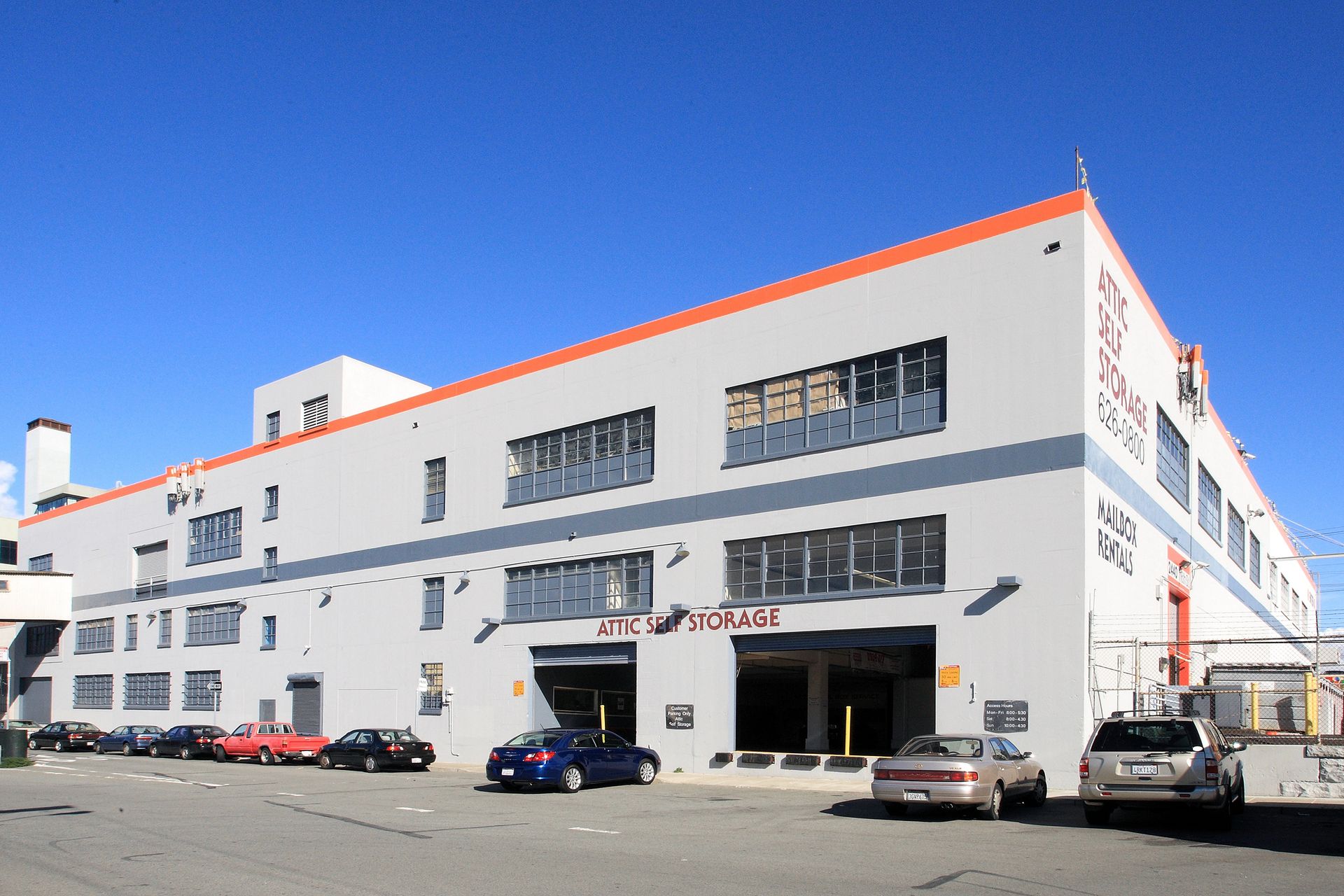 Multi-story industrial building, gray with red trim, blue sky, cars parked in front.