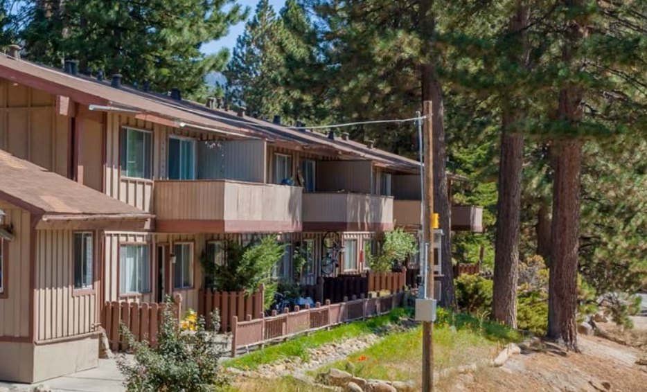 Brown and tan two-story townhouses with balconies, surrounded by tall pine trees, on a sunny hillside.
