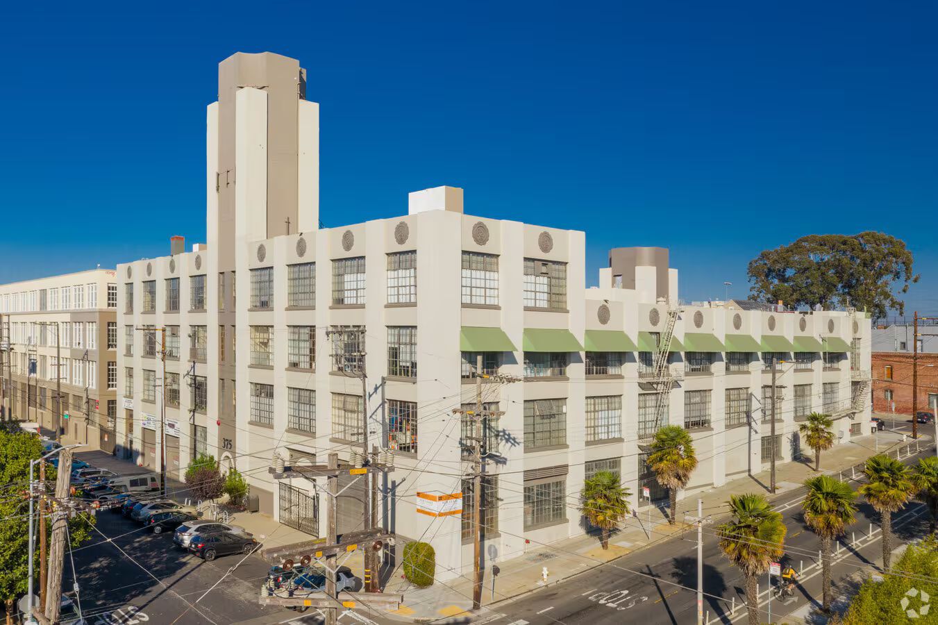 Multi-story, Art Deco-style building with a tower, light-colored walls, green awnings, and palm trees on a bright day.