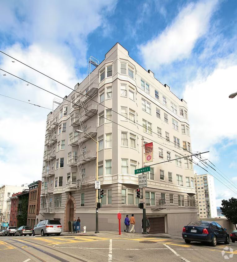 Multi-story apartment building at a San Francisco intersection with cars, pedestrians, and a blue sky.