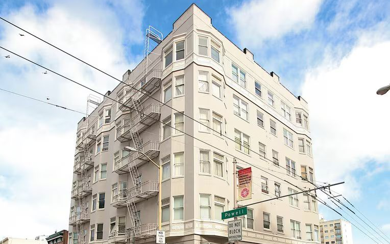 Multi-story beige apartment building on a street corner with a clear sky and power lines.