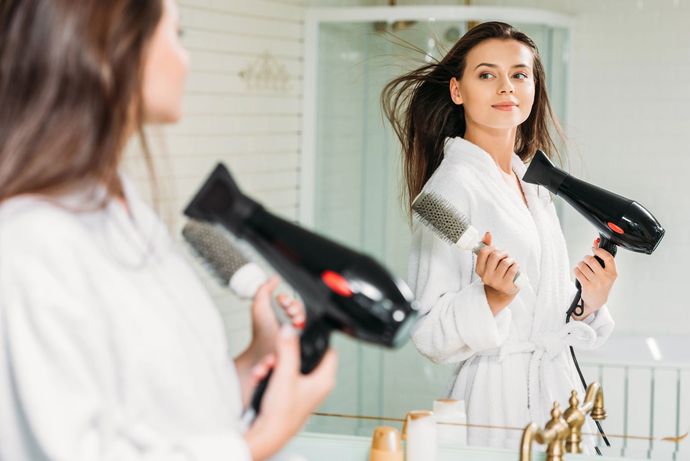 Una donna si asciuga i capelli con il phon davanti allo specchio del bagno, indossando un accappatoio bianco.