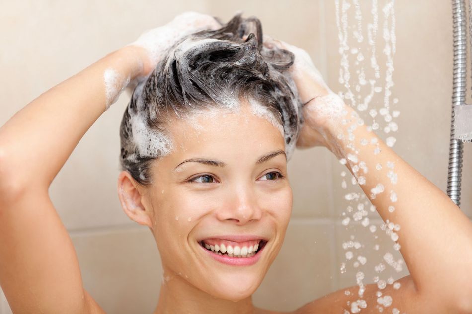 Una donna si lava i capelli sotto la doccia, sorridendo sotto l'acqua corrente e la schiuma dello shampoo.
