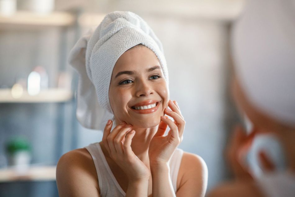 Una persona sorridente con i capelli avvolti in un asciugamano si applica dei prodotti per la cura della pelle davanti allo specchio del bagno.
