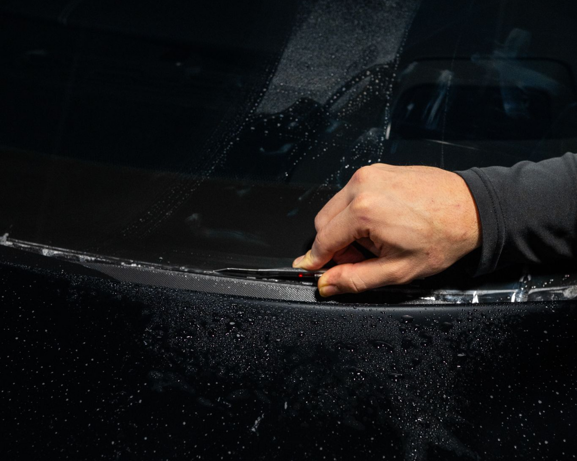 A close-up of a person's hand using a small tool to carefully apply a protective film to a wet, black car surface.