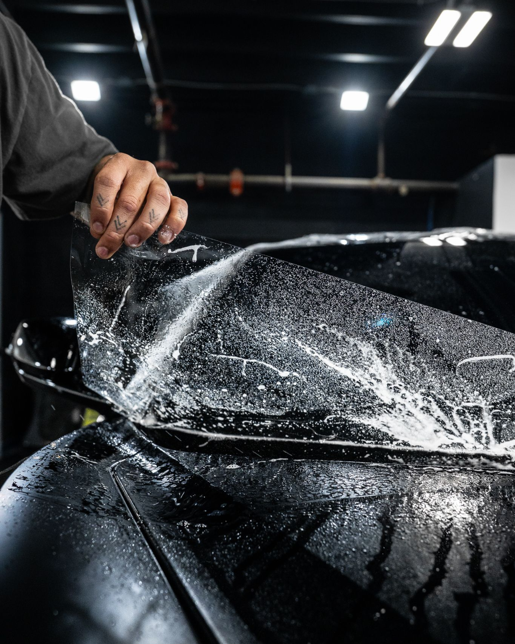 A hand applies a protective film to a car hood covered in soapy water, preparing it for a professional wrap installation.