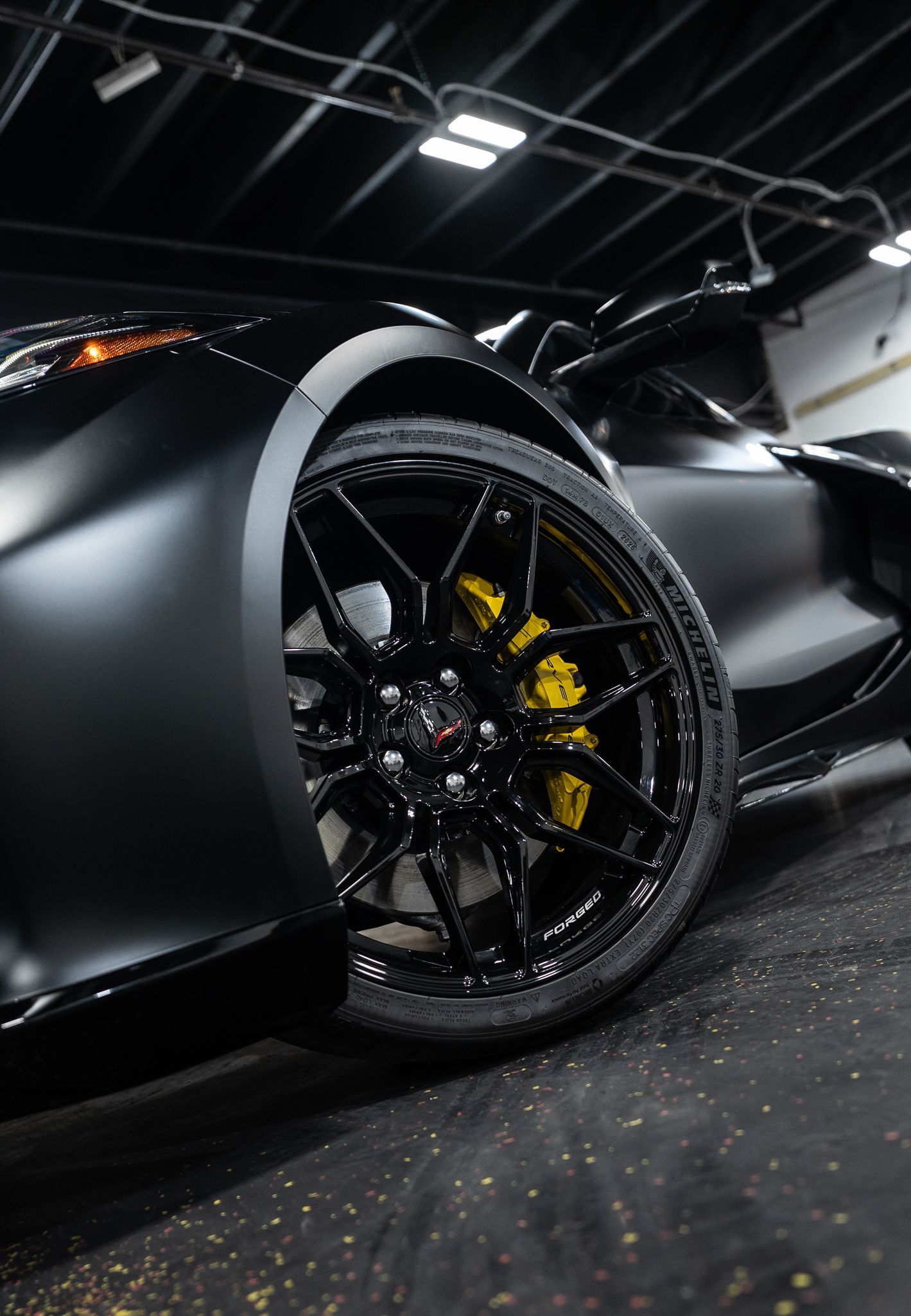 Close-up of a matte black sports car, highlighting its black wheel, yellow brake caliper, and sleek body lines.