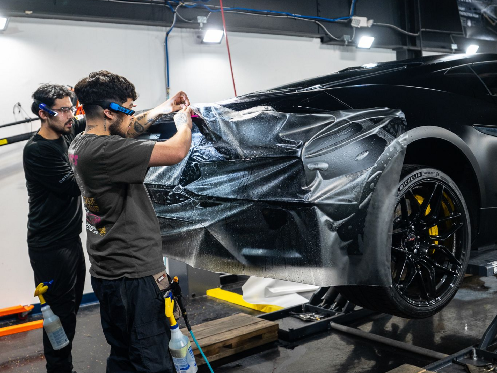 Two people applying film to a black car. One is holding a squeegee. Garage setting.