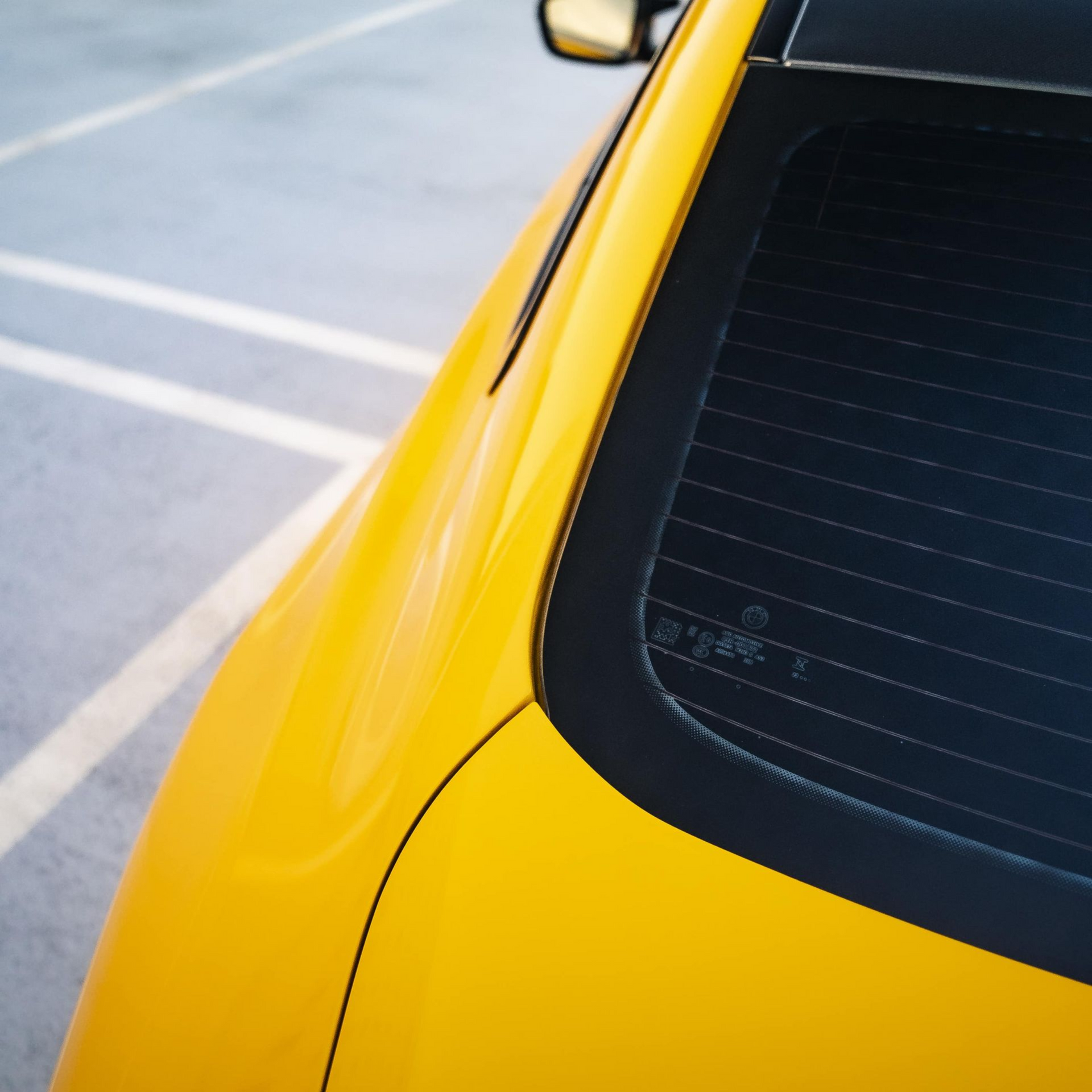 Yellow car's rear, angled view, showing window and body curves against a gray parking lot.