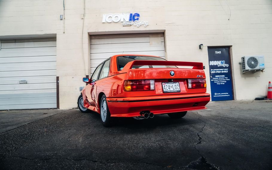 Orange BMW E30 sports car parked outside a garage with a black door and air conditioning unit.