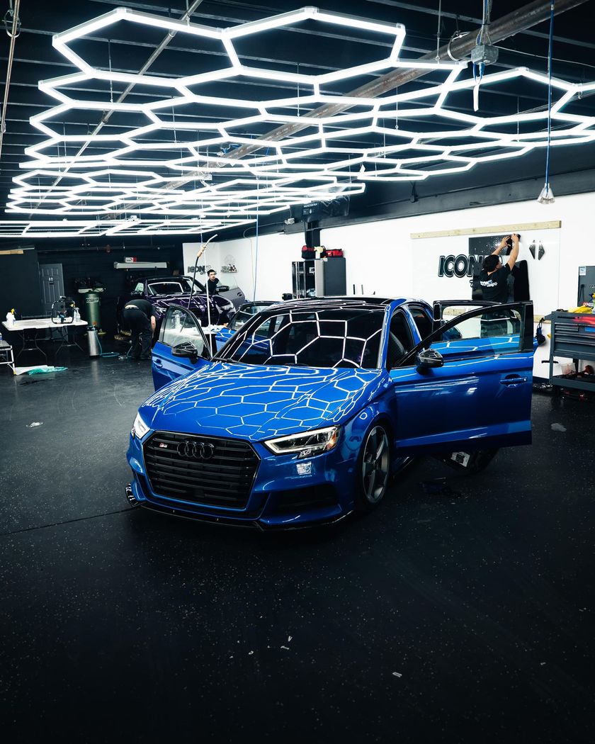 Blue Audi in a garage with honeycomb ceiling lights; doors open, a person standing.
