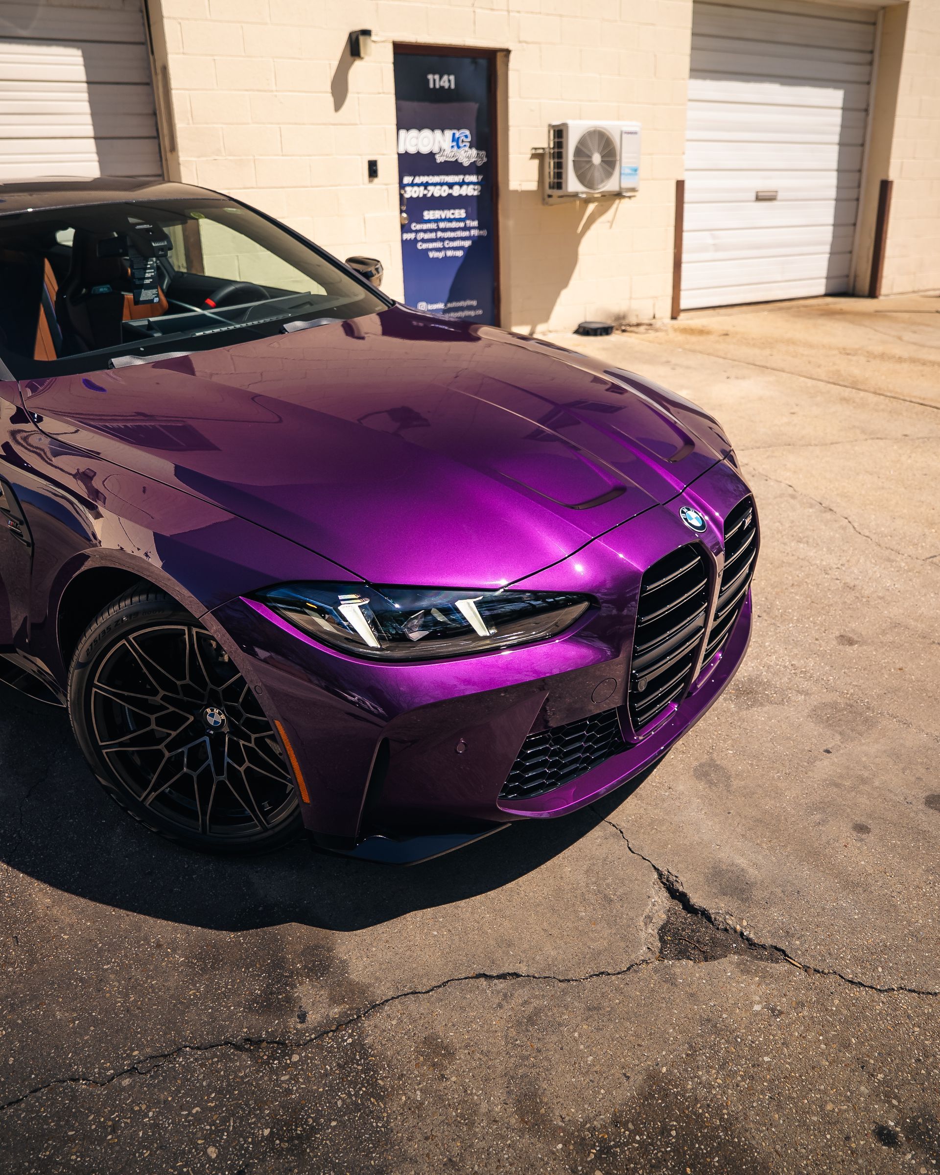 A purple BMW sports car parked outside an automotive shop with garage doors on a sunny day.