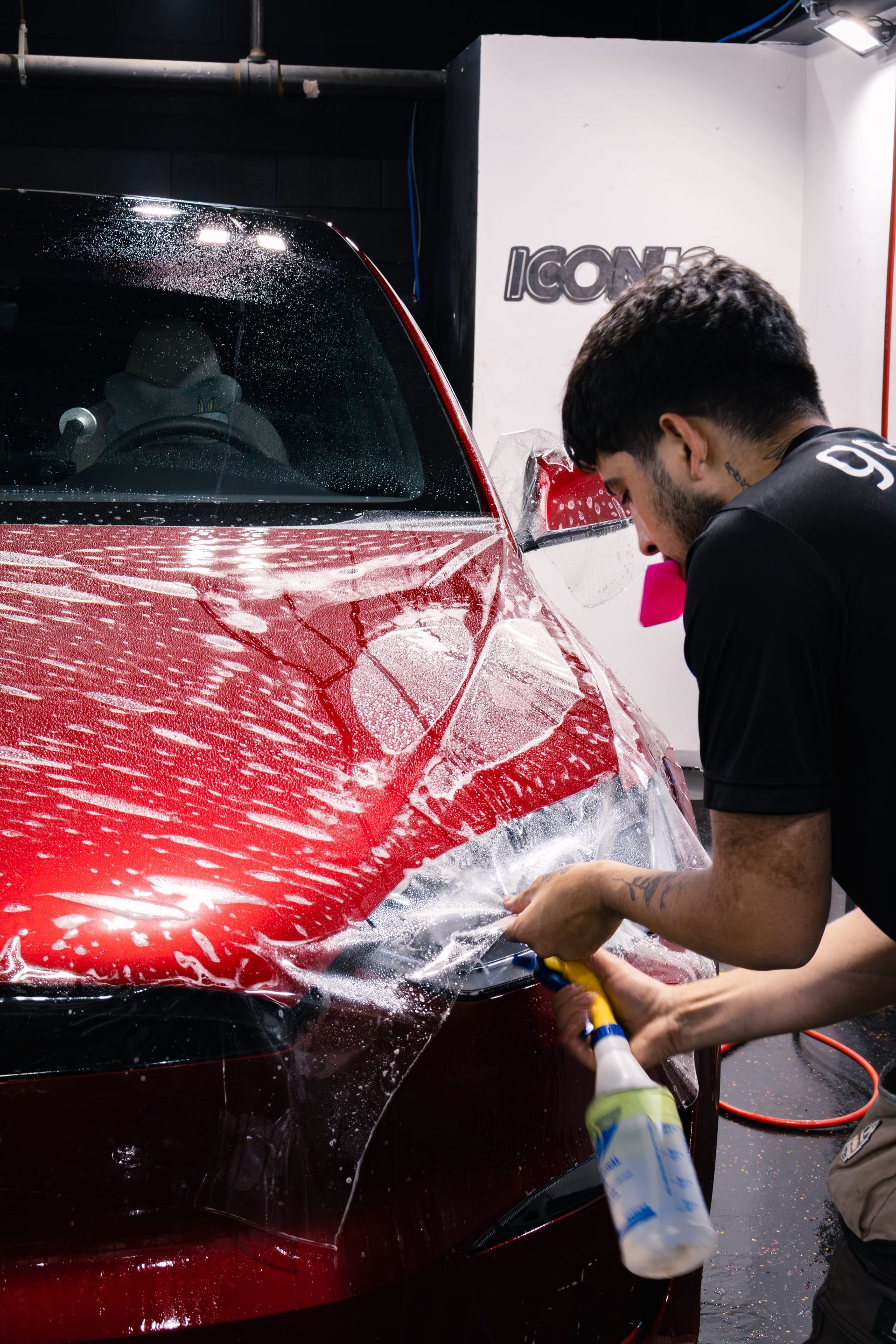 Person spraying solution on car surface, preparing to apply film. Indoor, workshop setting.