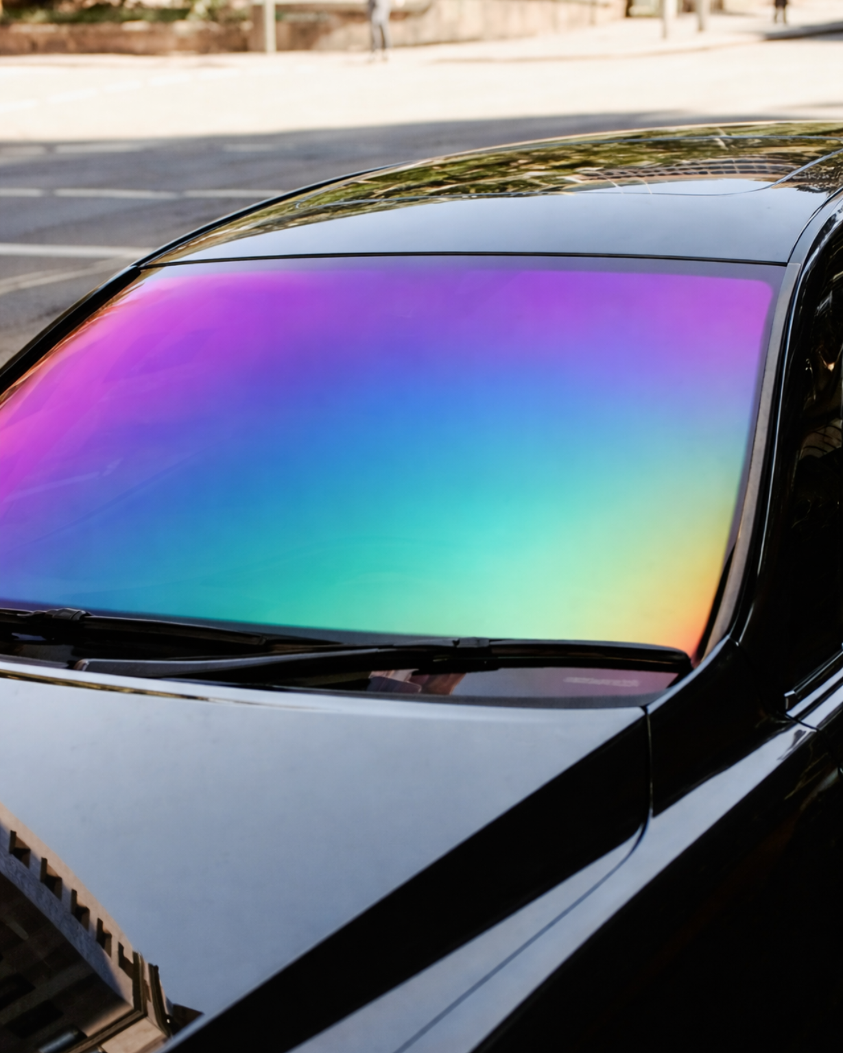 A black car with a windshield featuring a vibrant, iridescent rainbow film reflecting light.