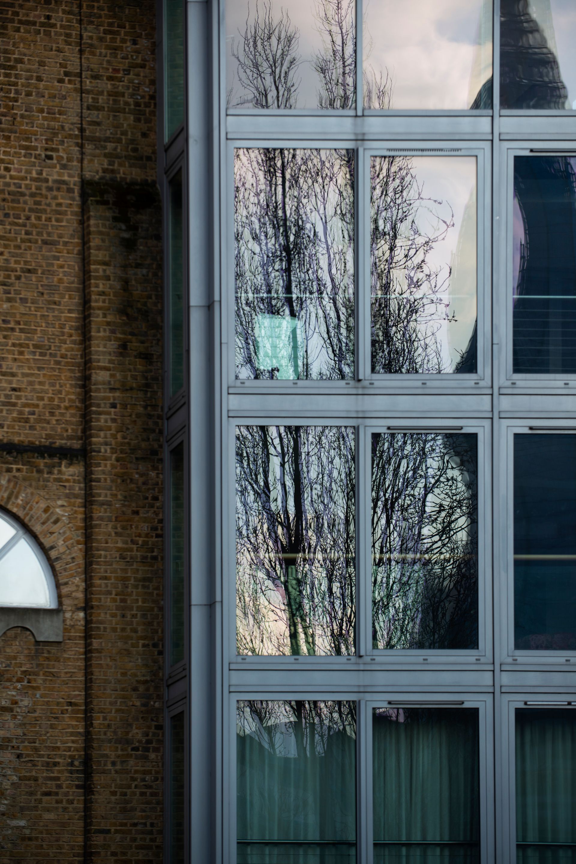 Brick wall beside a modern building with large reflective windows showing a tree's reflection and sky.