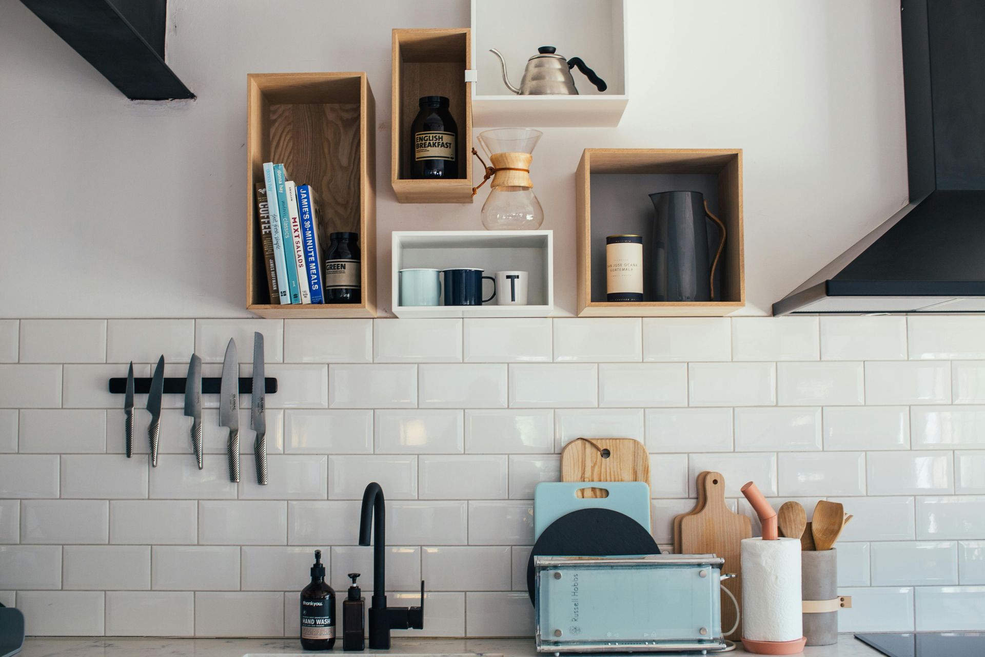 Kitchen interior with floating shelves holding coffee supplies and cookbooks above a tiled backsplash and sink.