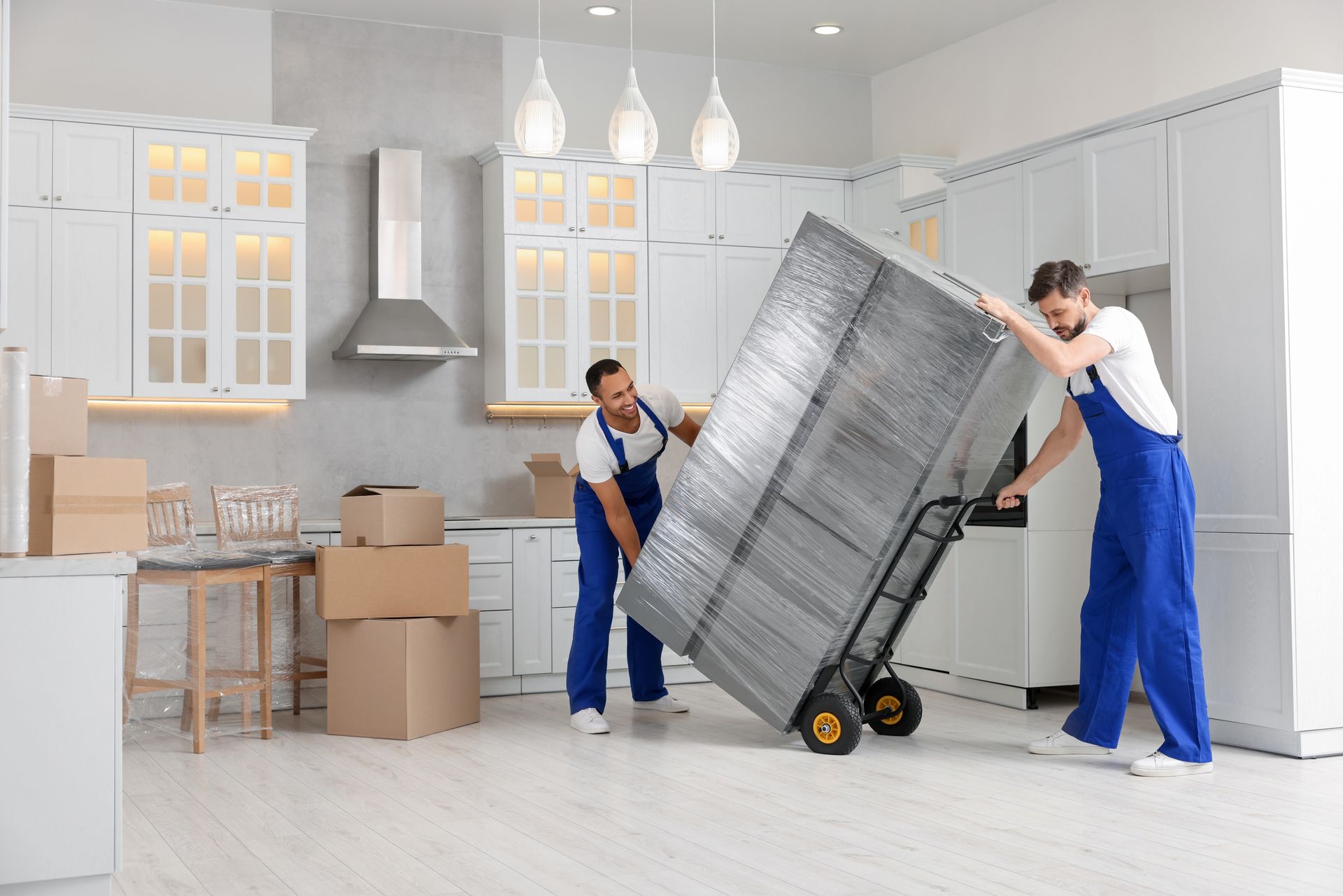 Two movers in blue overalls relocate a fridge in a modern kitchen with pendant lights. Two movers in blue overalls relocate a fridge in a modern kitchen with pendant lights.
