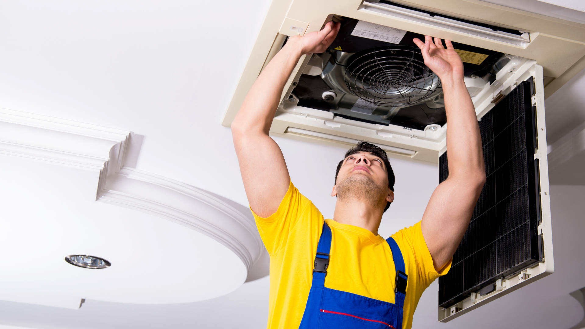 A man in a yellow shirt and blue overalls is working on an air conditioner.