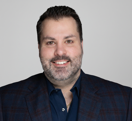 Man with dark hair and beard smiles, wearing a navy blue shirt and patterned blue blazer, in front of a gray backdrop.