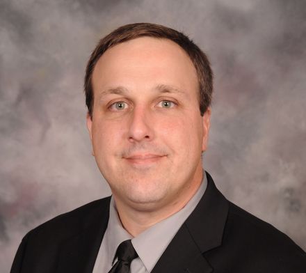 Man in a black suit and tie, smiling at the camera, against a white backdrop.