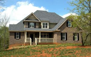 Two-story brick house with dark gray roof, front porch, black shutters, and light green surroundings.
