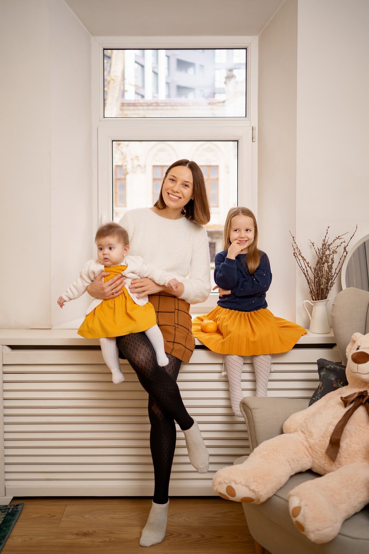 A woman is sitting on a window sill holding a baby and two girls.