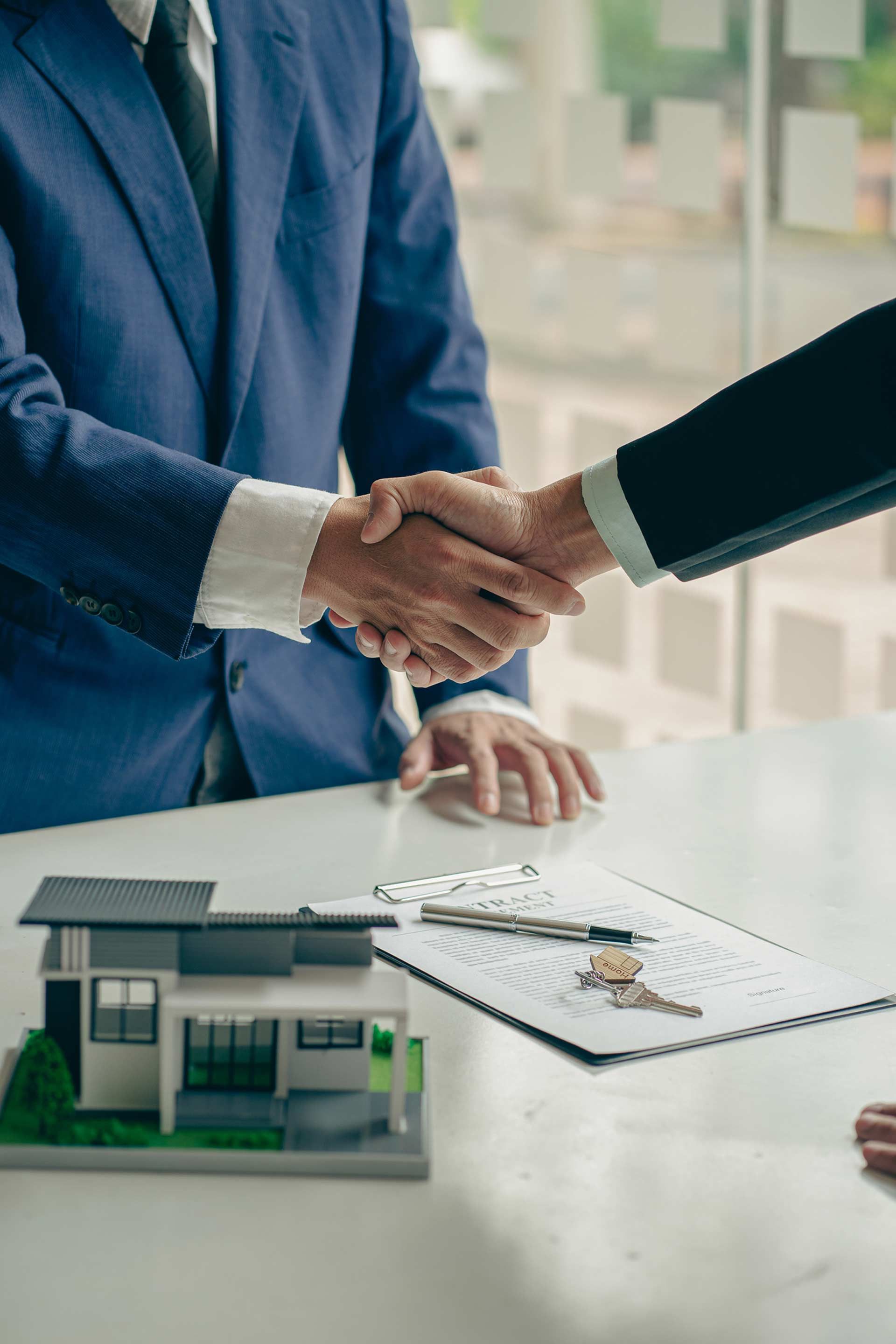 Two men are shaking hands over a table with a model house and keys.