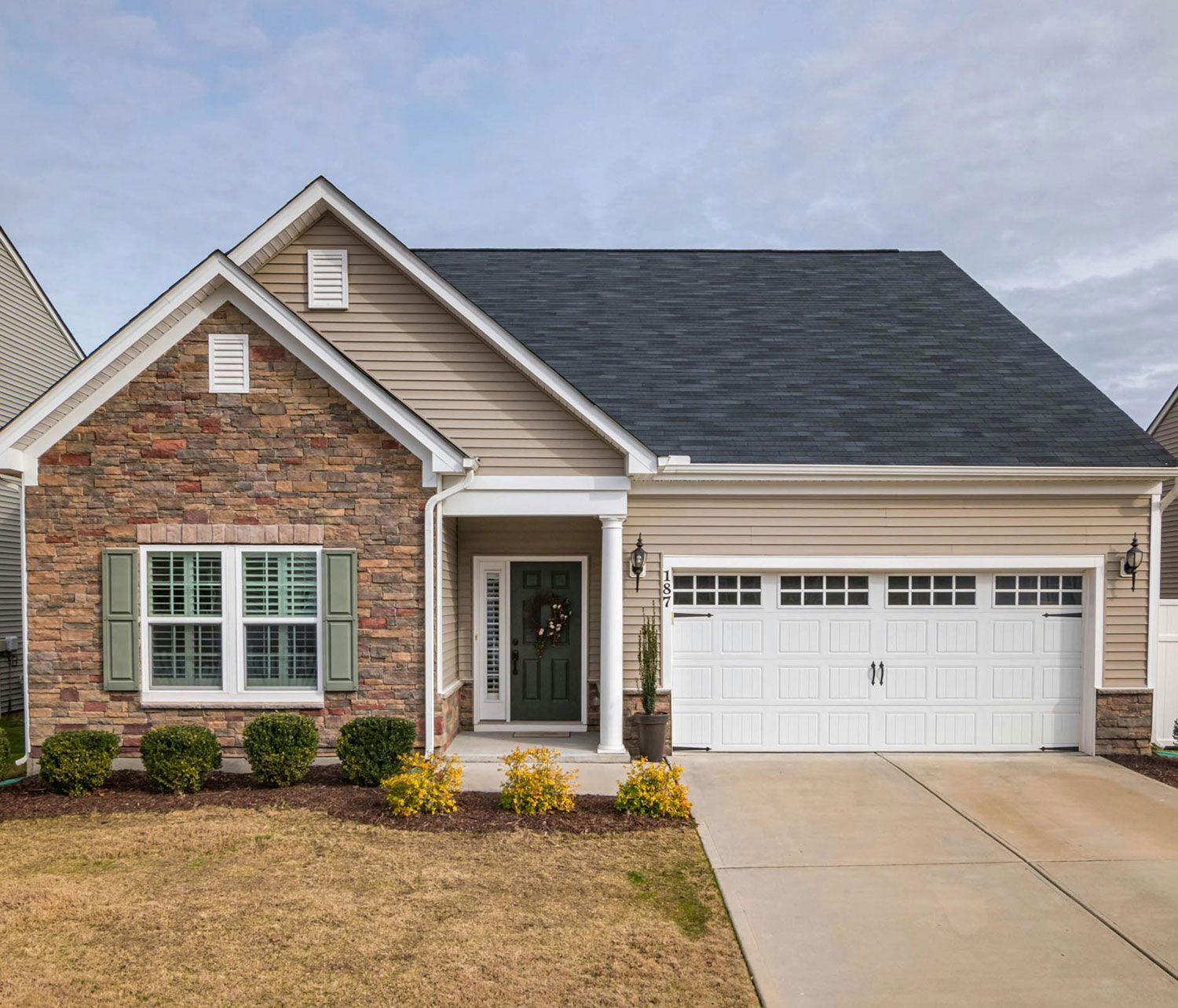 Tan and stone-faced house with a white garage door, black roof, and green shutters in front of a lawn.
