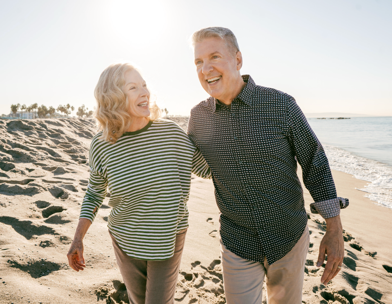 Couple walking and laughing on a beach, sunlit. Woman in stripes, man in patterned shirt.