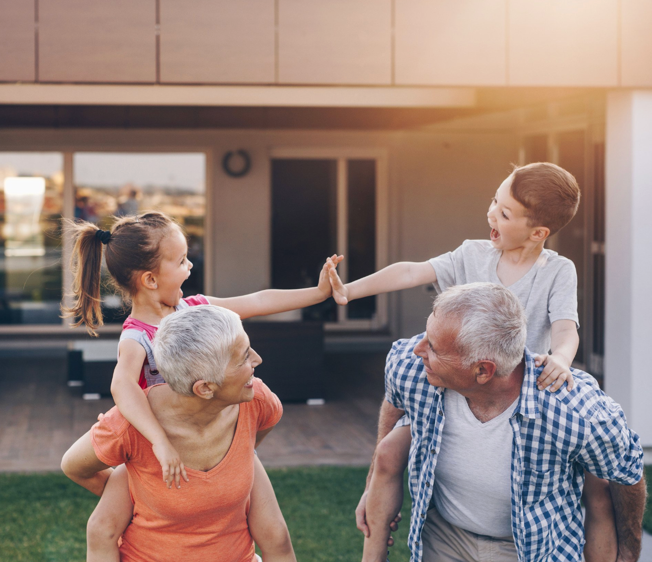 Grandparents giving piggyback rides to laughing grandchildren on a lawn.