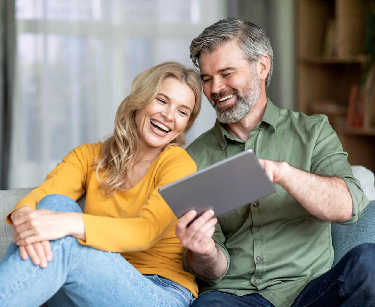 Couple laughing while looking at tablet on a couch; indoor setting.