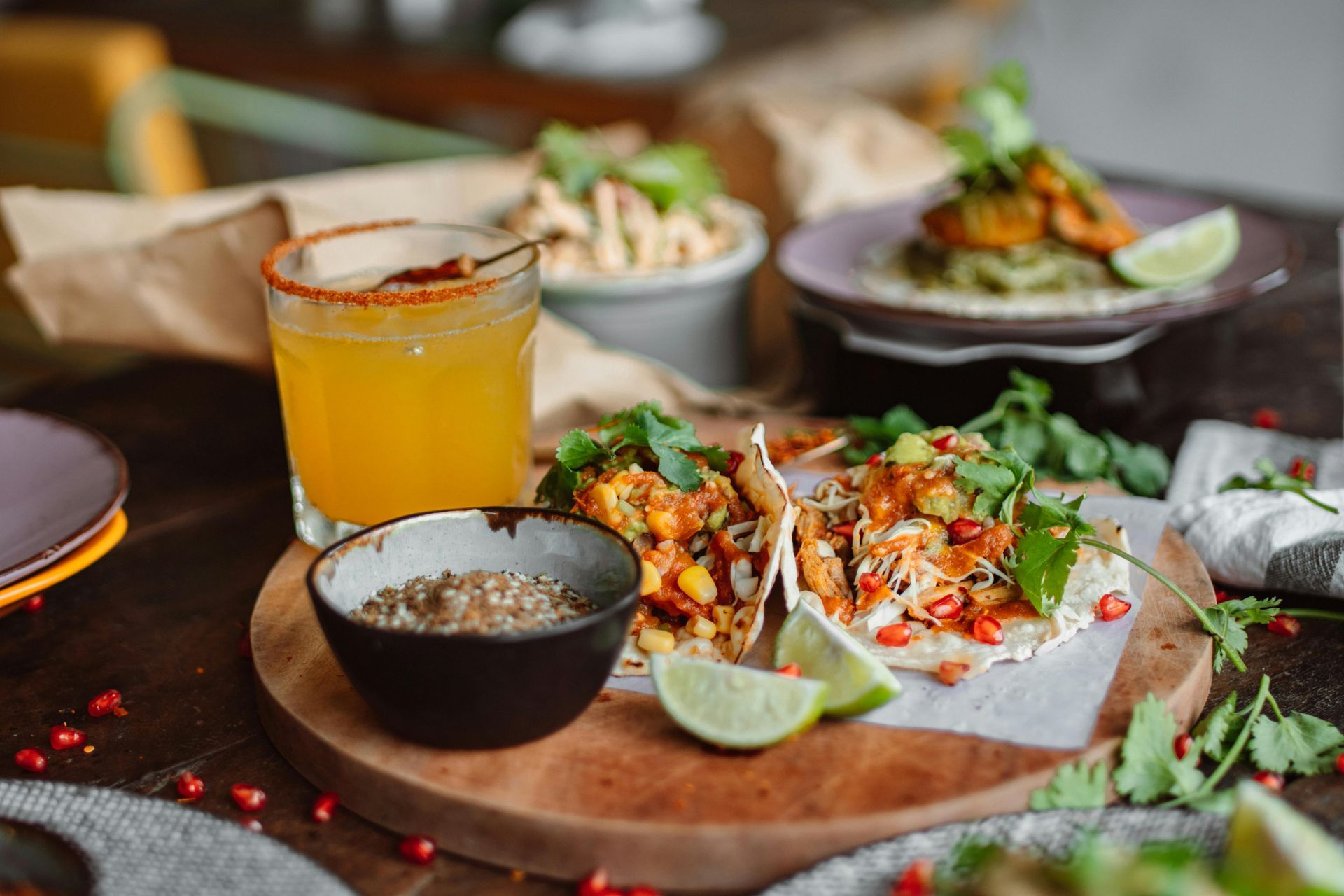 Tacos on a wooden board with a drink and a small bowl of spices. Other dishes and garnishes are also present.