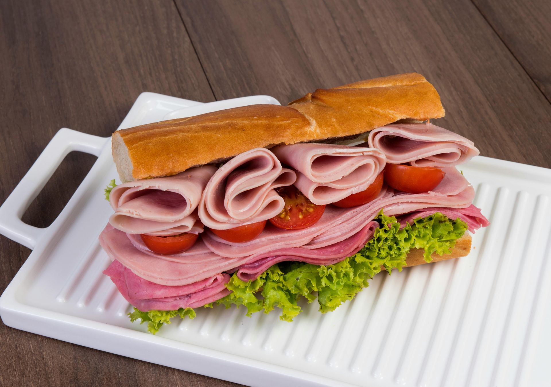 Sandwich on a white tray with ham, tomatoes, and lettuce on a baguette.