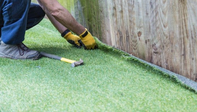A person wearing yellow gloves installs artificial turf along a wooden fence, using a hammer to secure the edge.