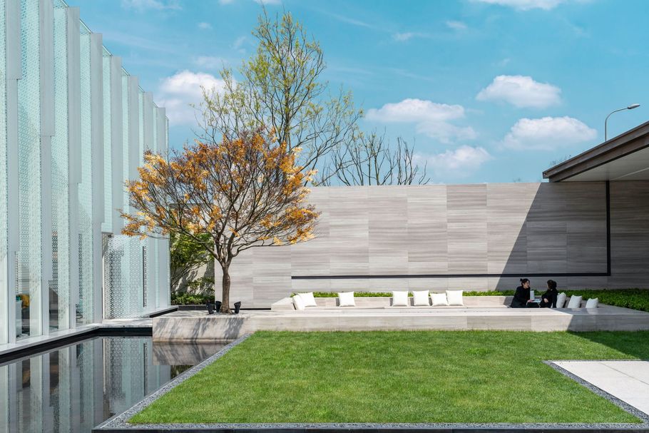 A modern outdoor courtyard featuring a reflecting pool, green grass, a small tree, and a stone seating area with cushions.